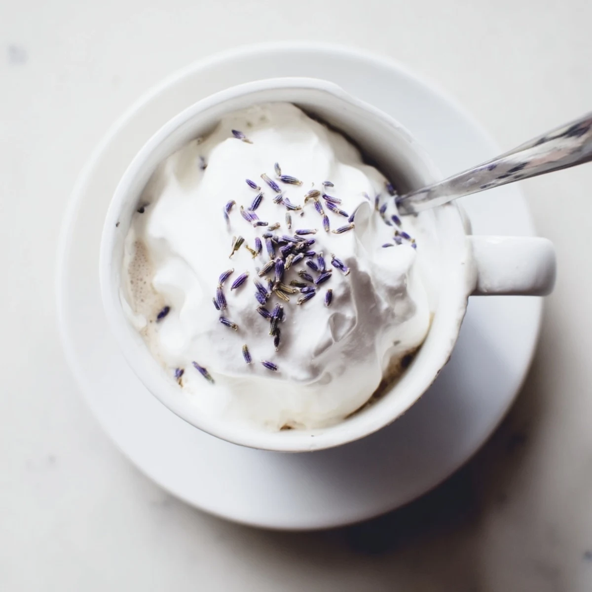 A close-up of a Whipped Lavender Latte in a ceramic mug, with cloud-like foam swirled atop creamy espresso and a few dried lavender buds garnishing the top.