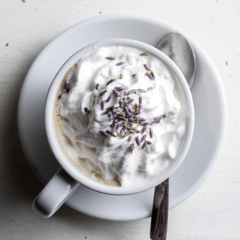 Two steaming mugs of Whipped Lavender Latte sit on a rustic wood table, showcasing velvety foam and a light dusting of lavender beside a scone.